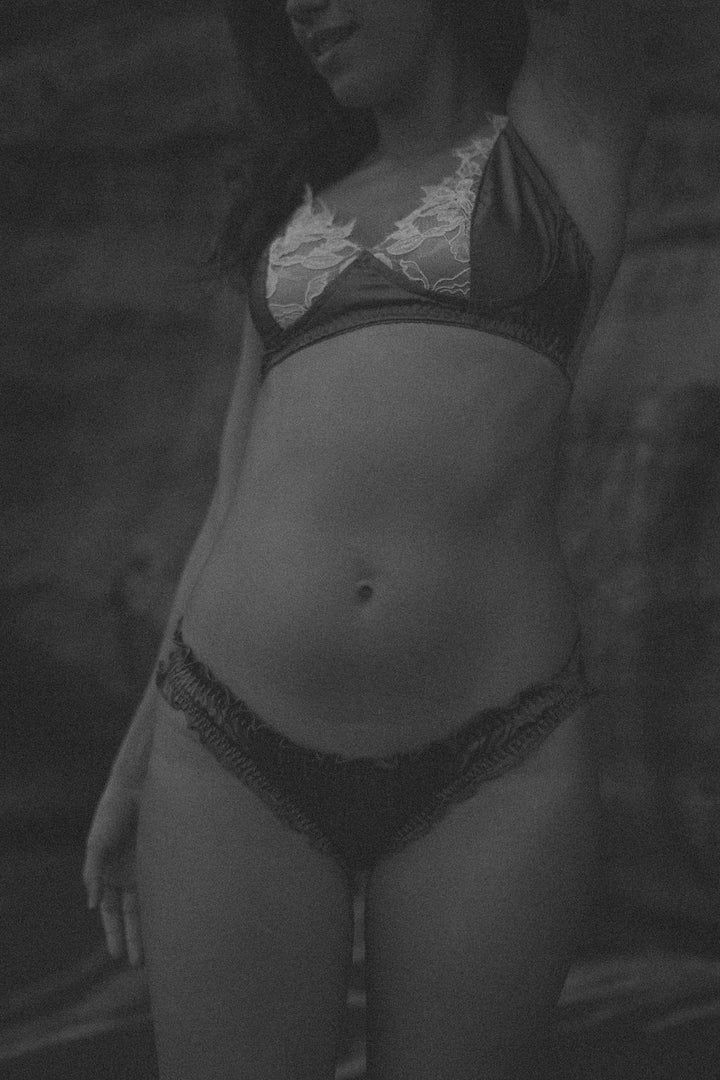 A black and white grainy photo of a woman posing in front of cliffs wearing luxury silk lingerie. She wears a pair of "Silk Mondays" Amie silk ruffle panties and a matching Amie silk bralette. Her face is turned from the camera
